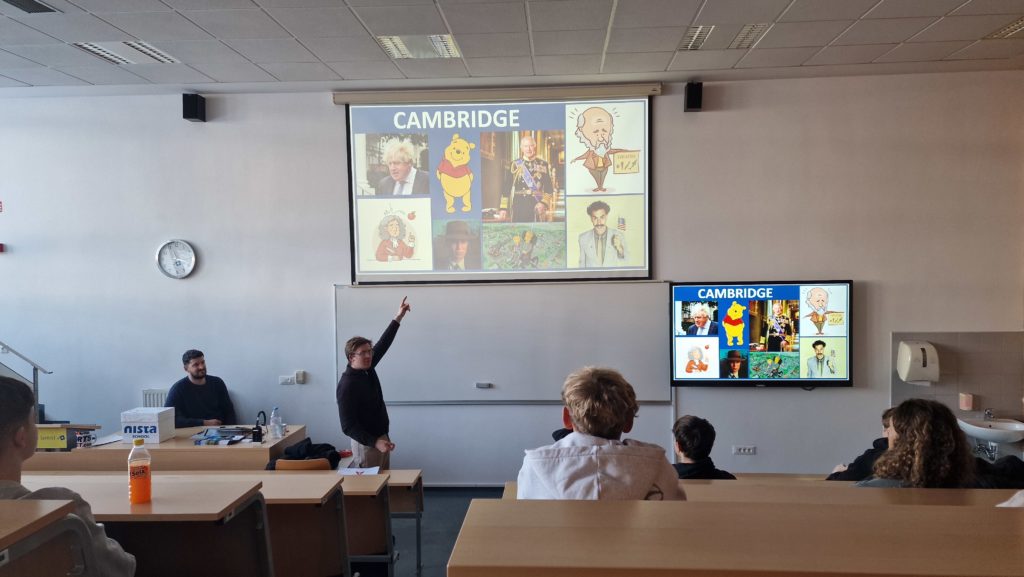 Predstavitev v učilnici na Gimnaziji Šentvid. Predavatelj stoji pred belo tablo in z dvignjeno roko kaže proti prosojnici z naslovom Cambridge, ki prikazuje kolaž ilustracij in znanih simbolov, vključno z medvedkom iz risanke. Dijaki sedijo v klopeh in spremljajo predstavitev. Ob tabli je dodatni monitor s prikazom iste prosojnice, v ozadju pa sta vidna še zvočnika in šolska učilnica.