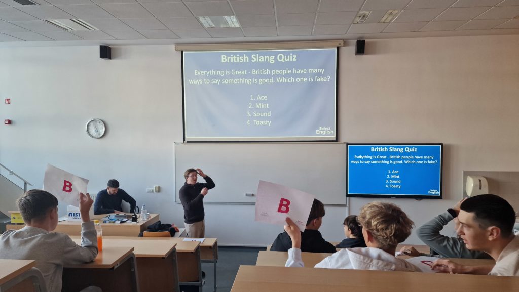 Učilnica na Gimnaziji Šentvid med interaktivnim kvizom British Slang Quiz. Predavatelj stoji pred belo tablo in projekcijskim zaslonom z vprašanjem kviza, dijaki pa sedijo v klopeh in dvigujejo liste z odgovorom ‘B’. Del predstavitve je prikazan tudi na stranskem monitorju. V učilnici so vidni šolske klopi, stenska ura, zvočniki in sproščeno, aktivno vzdušje.