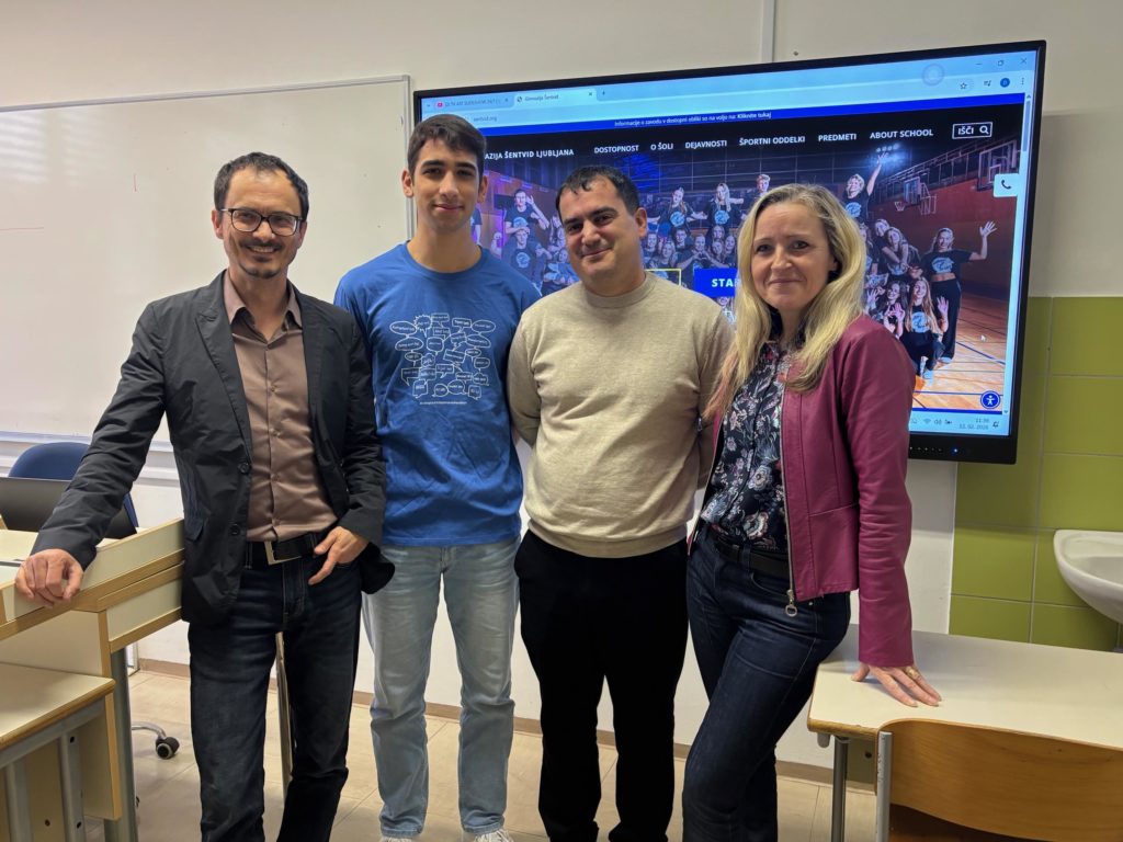 Zmagovalec ponosno stoji pred tablo v družbi treh profesorjev. V ozadju zaslon, na njem je prikazana spletna stran šole.