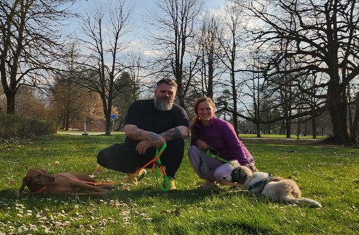 Petar Babić in Ula Brank Pečko klečita na travniku v parku na sončen dan, obdana z drevesi brez listja. Vsaka oseba drži psa na povodcu. Na levi strani leži pes z rjavo dlako, na desni pa pes s svetlo in temno lisasto dlako, ki leži na travi. Oba psa sta sproščena, prizor pa deluje kot umirjena vadba ali druženje z živalmi na prostem.